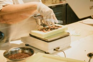 Chef in gloves arranging chocolate and ingredients on a kitchen scale for confectionery preparation.